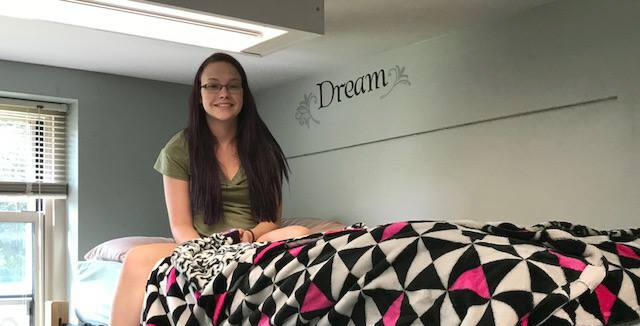 Image alt. text: photo of sophomore Alex on a lofted dorm room bed with a hot pink, black, and white bedspread. She is smiling and looks a little terrified.